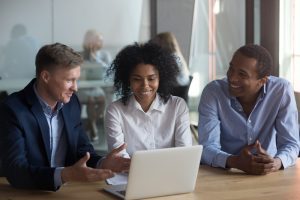 Positive Black Woman Sitting At Desk With Diverse Male Colleagues